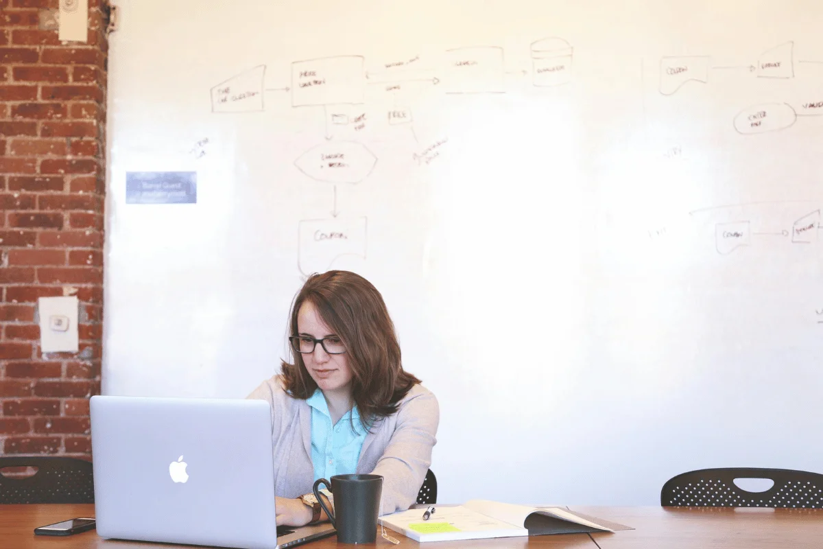 Person working on a laptop at a table with a black mug, notebook, and phone, in front of a whiteboard with diagrams and a brick wall.