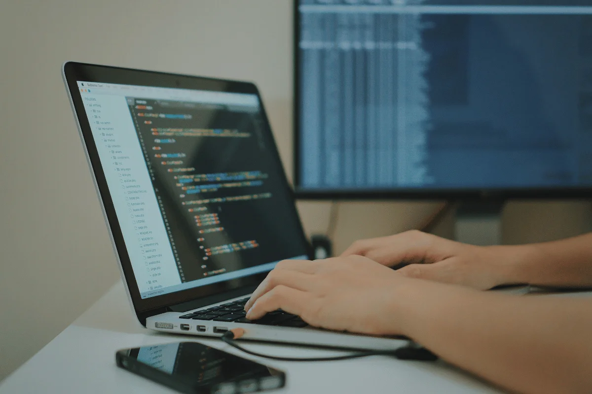 Programmer typing on a laptop with code on screen and an external monitor in the background.