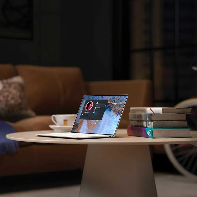 Thin laptop on white round table next to stacked books and a coffee cup with a blurred brown couch in the background.