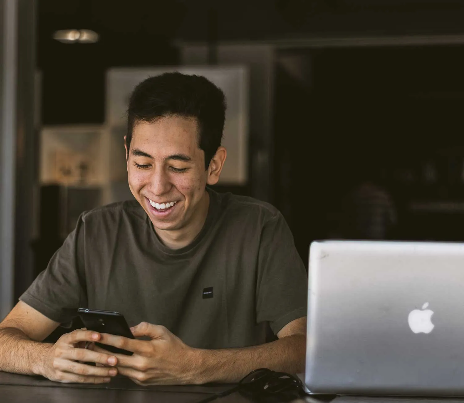 Smiling young man looking at his smartphone while sitting at a table with a silver Apple laptop.
