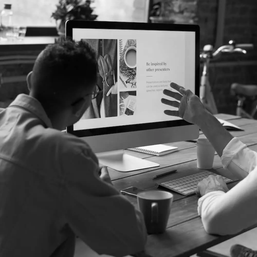 Two people sitting at a wooden desk looking at a large computer screen displaying images and text about being inspired by other presenters.