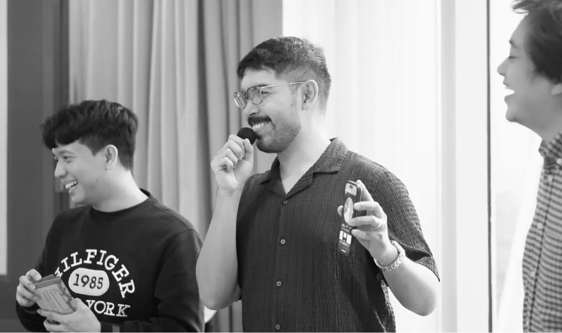 Three men smiling and laughing indoors, one holding a microphone and a phone, another holding a notebook.