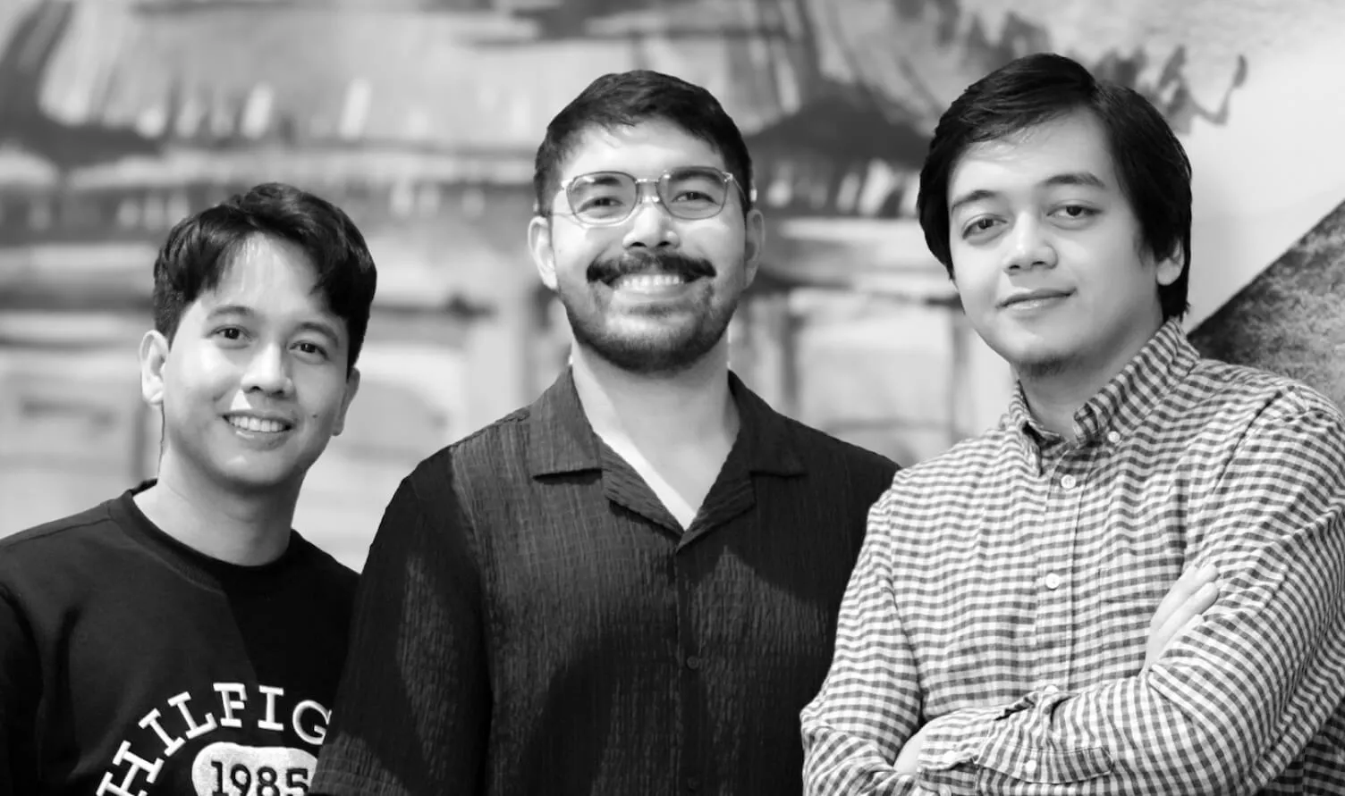 Black-and-white photo of three men smiling at the camera, one wearing glasses and a dark shirt, another in a dark sweatshirt, and the third in a checkered shirt with arms crossed.