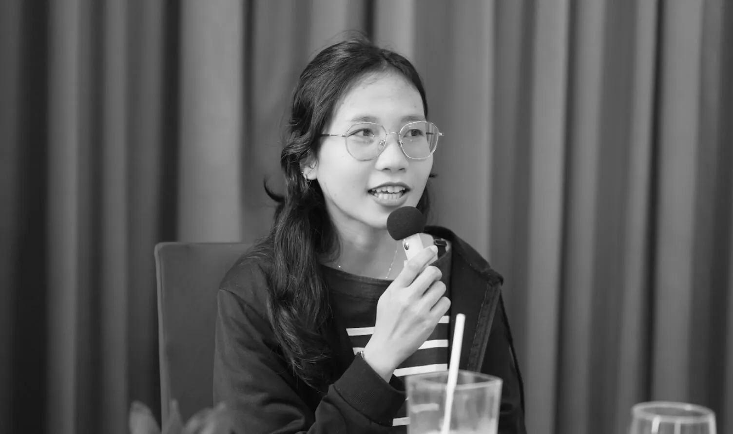 Woman with glasses and long hair speaking into a microphone while sitting at a table with a glass and straw in front of her.