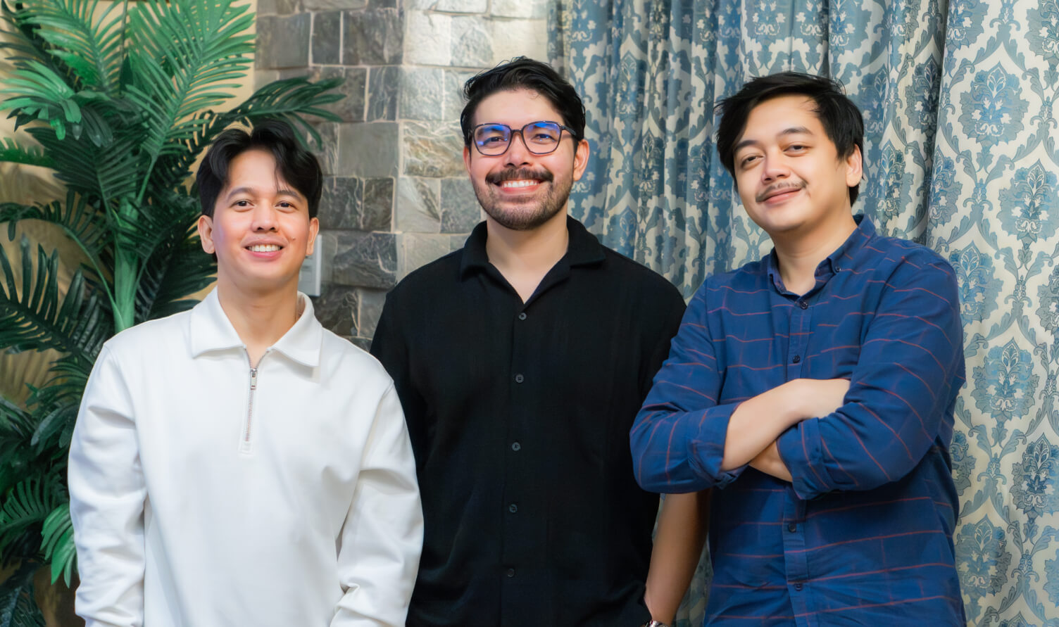 Three men standing indoors, smiling, with one wearing a white zip-up shirt, another in a black button-up shirt and glasses, and the third in a blue checkered shirt with arms crossed.