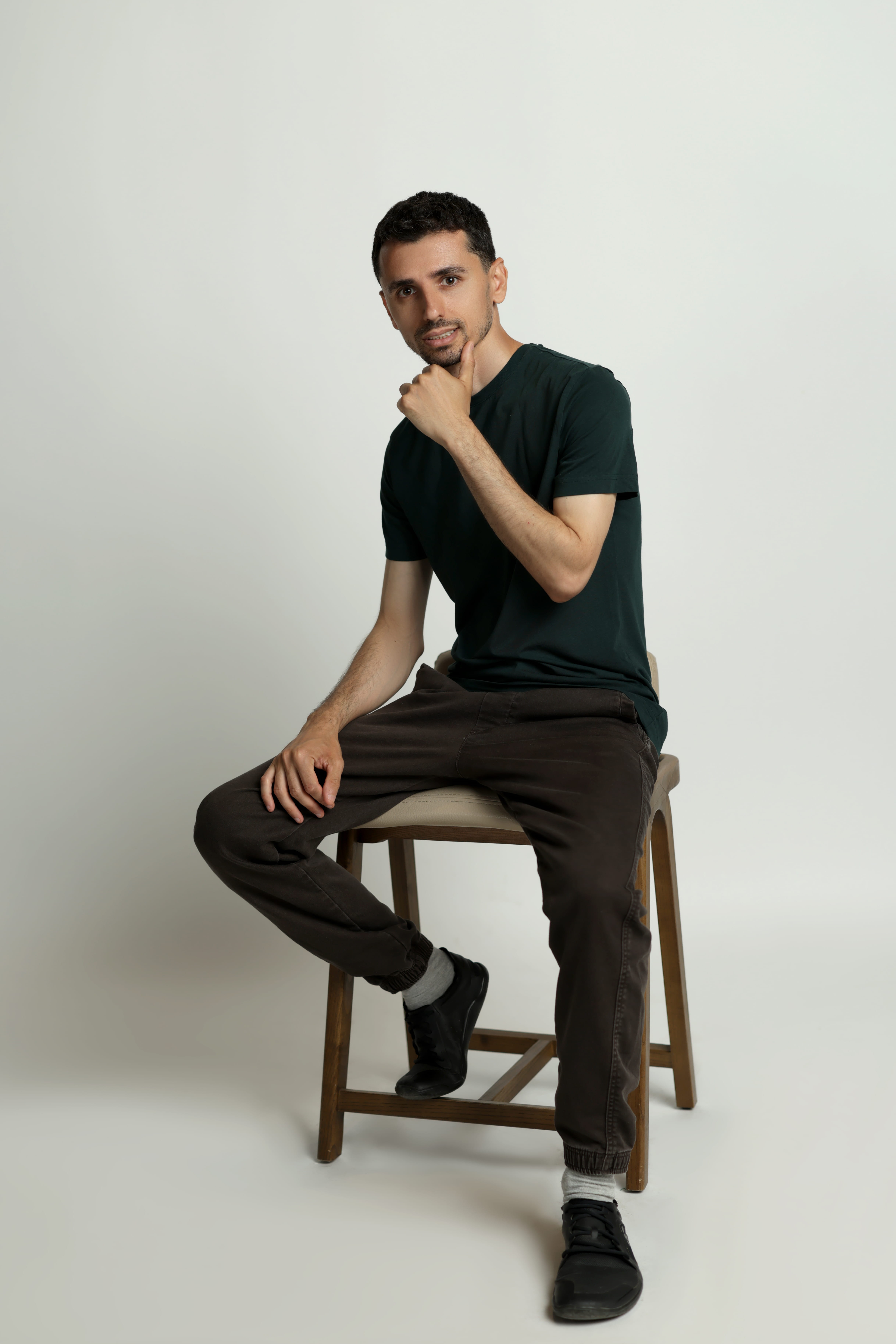 Man sitting on a wooden stool wearing a dark green shirt, brown pants, and black shoes.