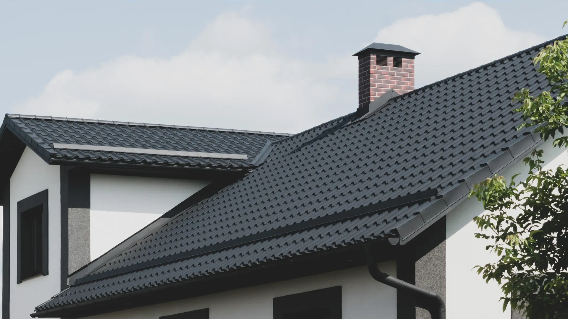 Modern house with a black metal roof, brick chimney, and surrounding greenery under a cloudy sky.