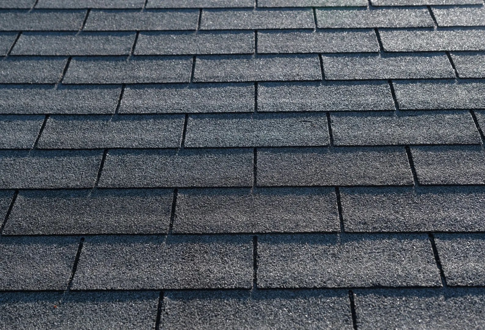 Close-up of gray asphalt roof shingles in sunlight, highlighting texture and pattern.