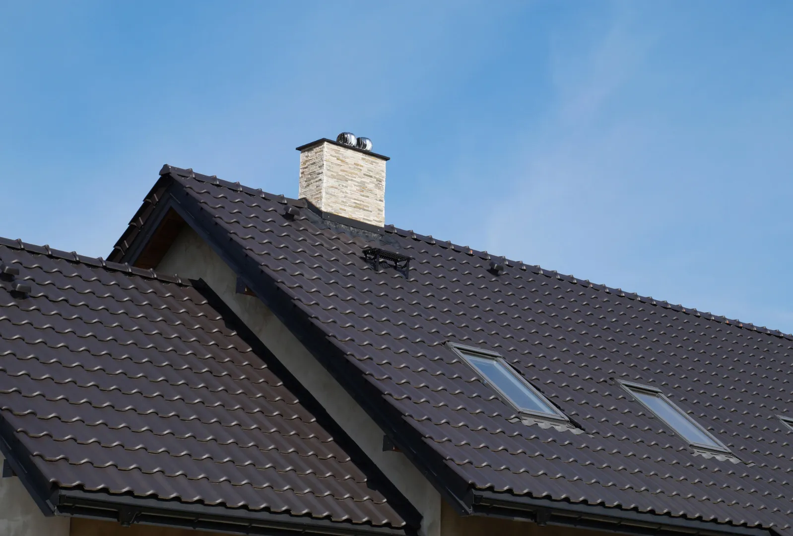 Modern house with dark tiled roof, skylights, and chimney against a clear blue sky.