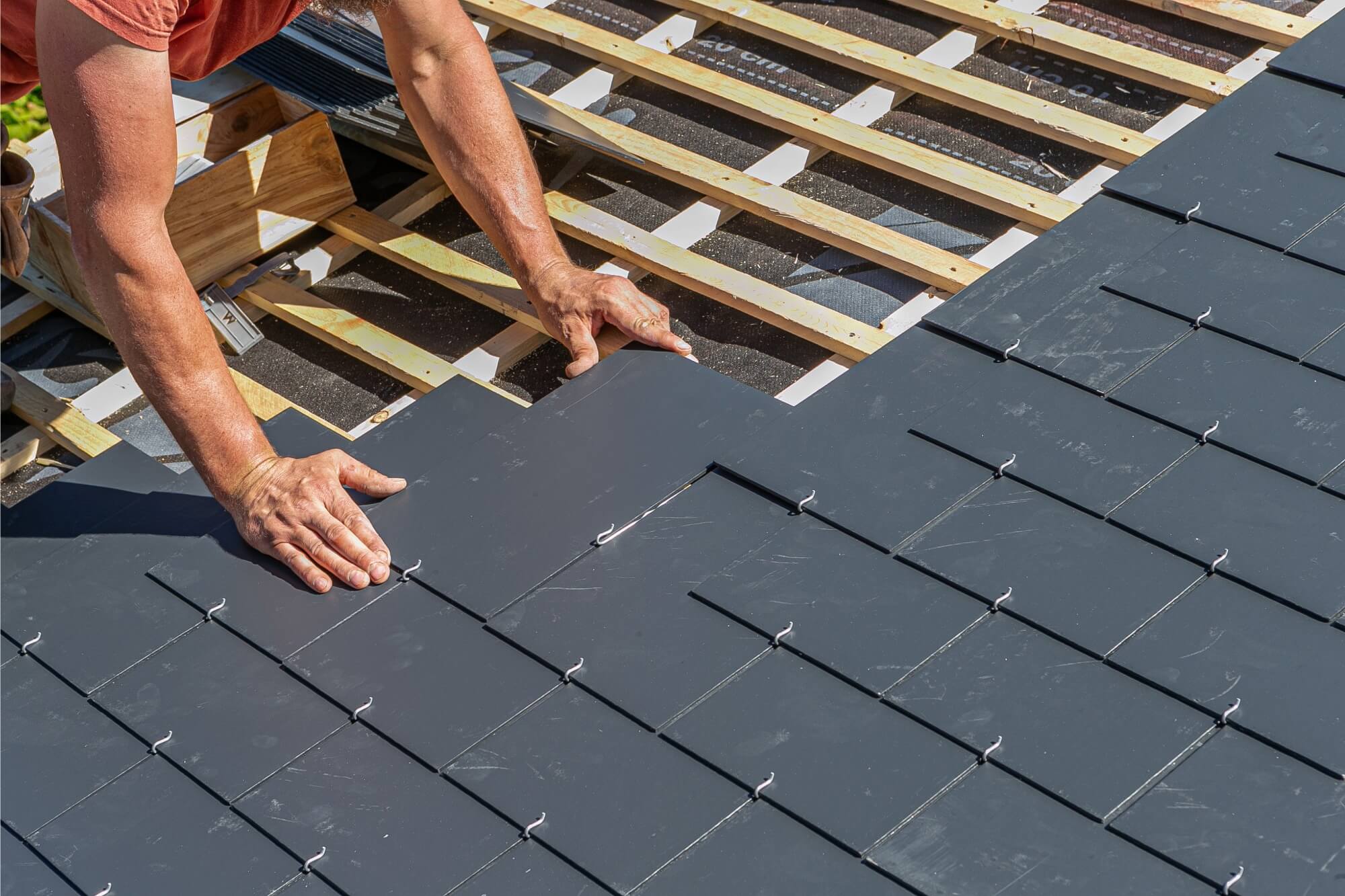 Worker installing dark slate tiles on a roof with wooden battens visible underneath.
