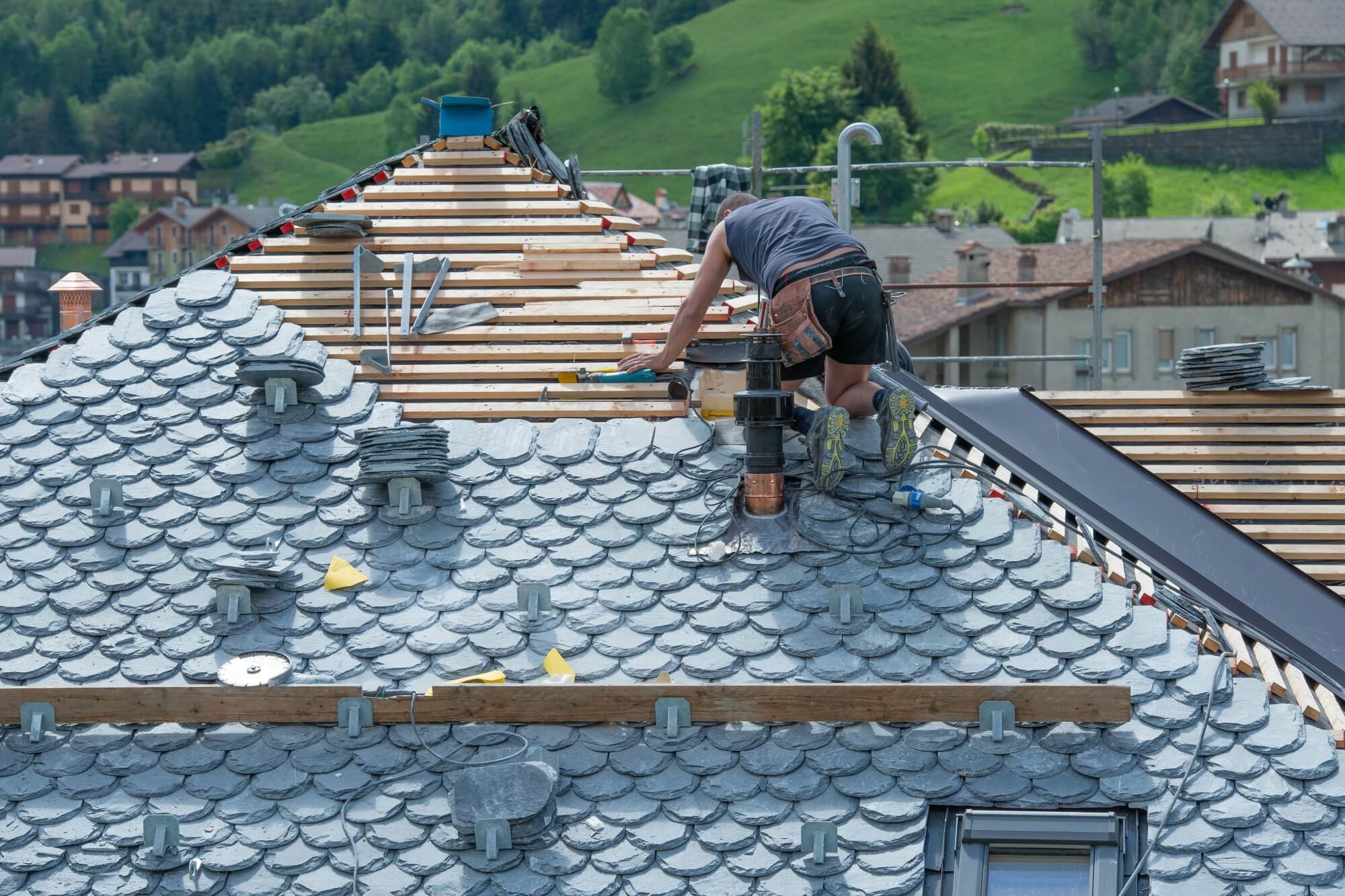 Worker repairing a slate roof on a residential building in a rural area with green hills.