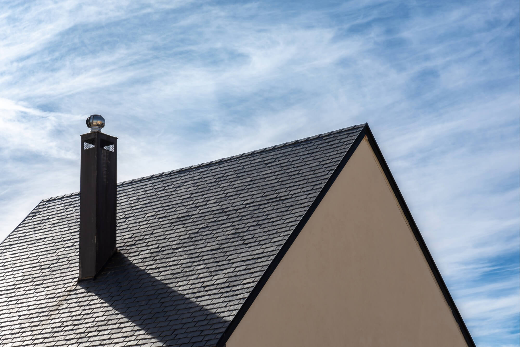 Modern house with a slate roof and chimney against a blue, cloudy sky background.