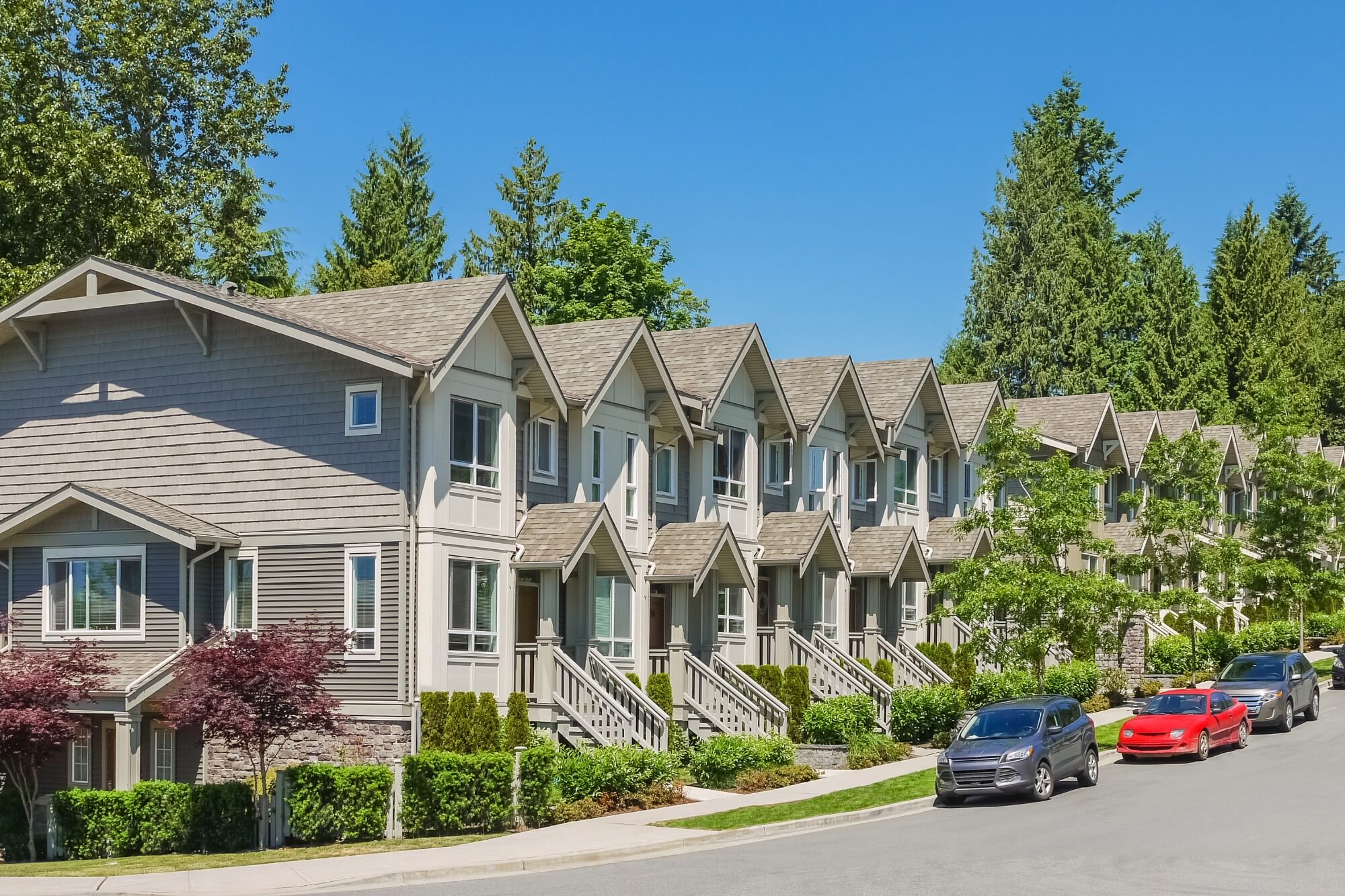 Row of modern townhouses with cars parked on the street, surrounded by lush green trees.
