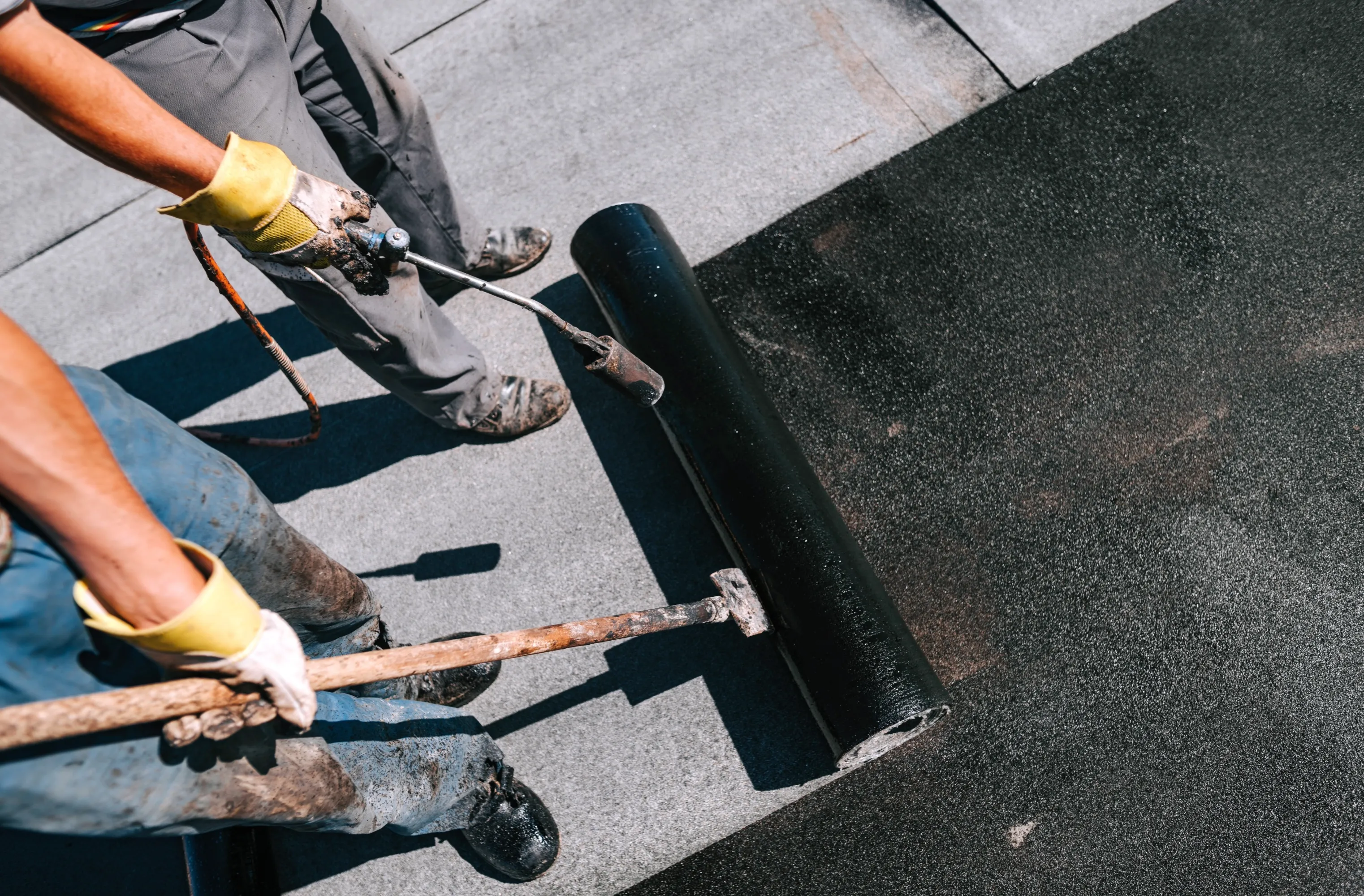 Workers applying black waterproofing membrane with torch on a flat roof for weatherproofing.