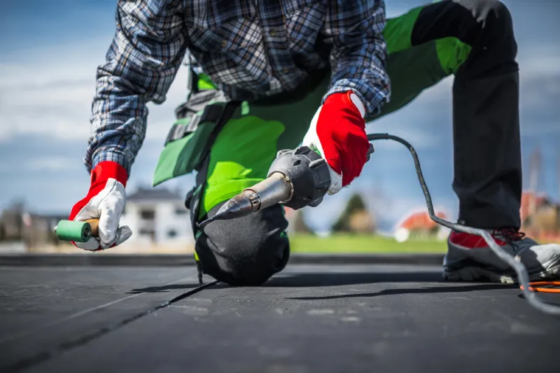 Worker wearing red gloves using a heat welding tool to seal a black EPDM roofing membrane.