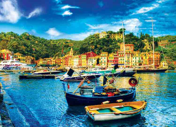 Boats in blue water with brown buildings on the coast and a green hillside with a blue sky