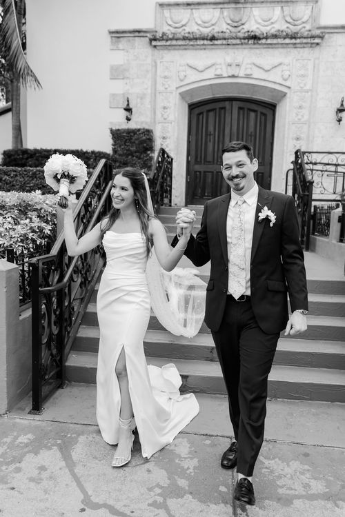 Black and white photo of a bride and groom joyfully walking out of a church, holding hands and smiling.