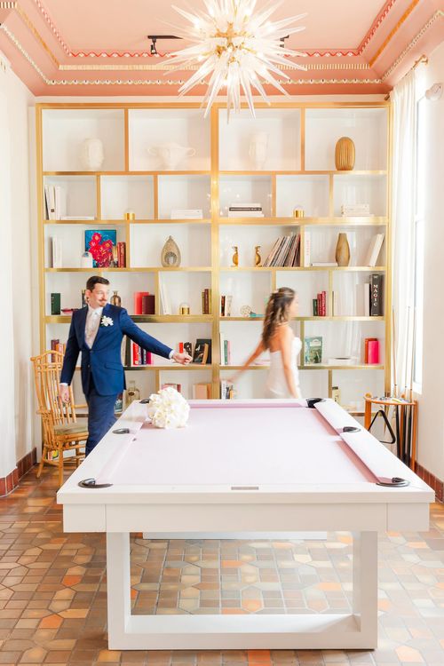"Bride and groom near ping pong table in a brightly lit room with elegant decor and a shelved wall