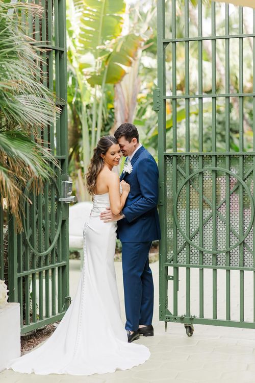 Bride and groom embracing in a romantic setting, surrounded by lush greenery and framed by a decorative metal gate.