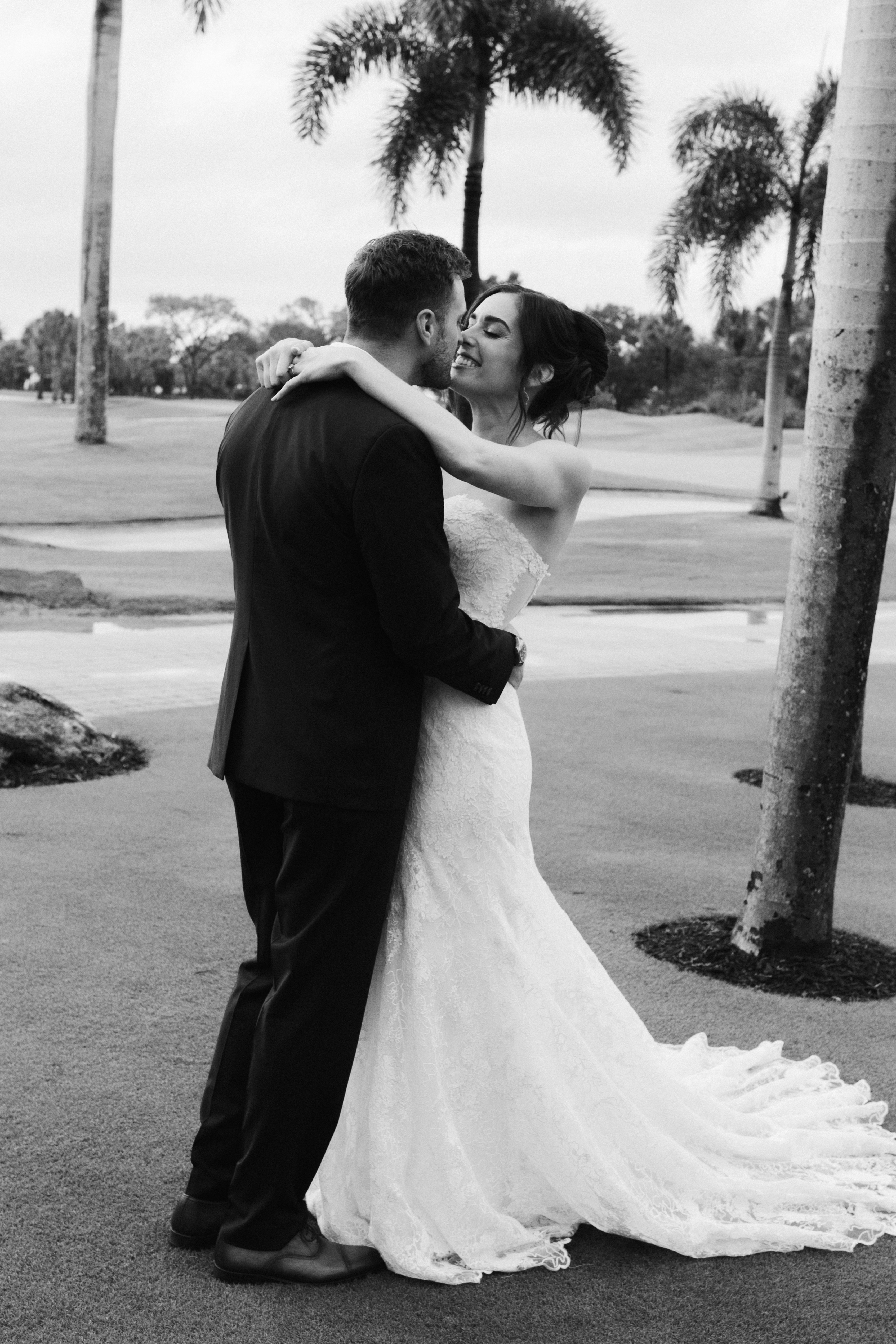 Black-and-white portrait of the bride and groom embracing beneath palm trees.