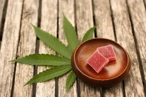 A small clay bowl with two pink sugar coated gummies, a cannabis leaf underneath, all on top of a slotted aged wooden surface