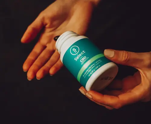 A soft and warm lit pair of hands, one holding a bottle of Select CBD capsules and the other open as a capsule comes out of the bottle