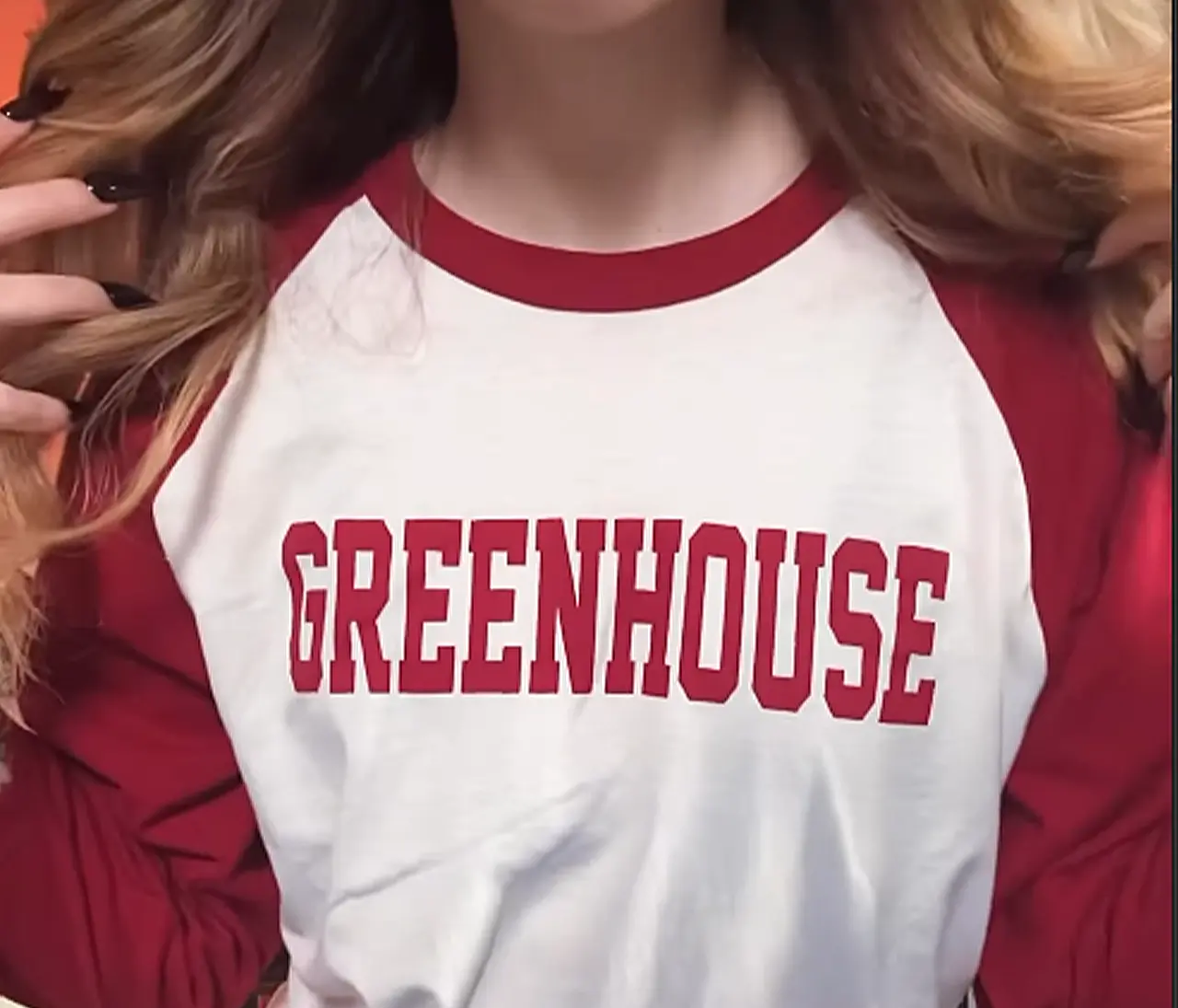 Gemini said A close-up photograph of a model wearing a two-tone baseball-style tee with a white body, maroon long sleeves, and a matching maroon neckline. Across the chest, the word "GREENHOUSE" is printed in large, bold, maroon block letters with a slightly curved arch. The model, whose face is partially obscured by her long blonde hair and dark manicure, holds the collar of the shirt open.