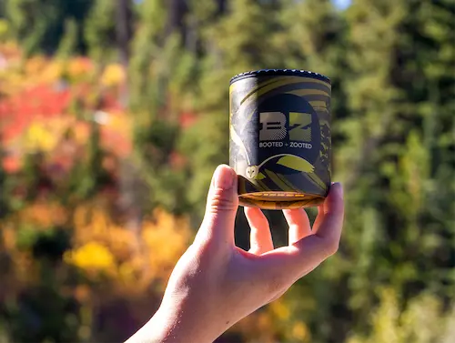 A hand holding a container of Booted + Zooted cured rosin infused gummies on a sunny day in front of a blurred fall forest background