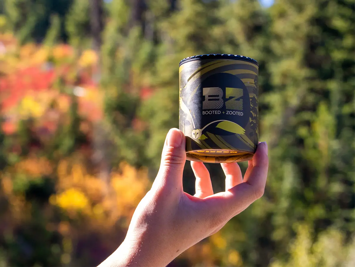 A hand holding a container of Booted + Zooted cured rosin infused gummies on a sunny day in front of a blurred fall forest background