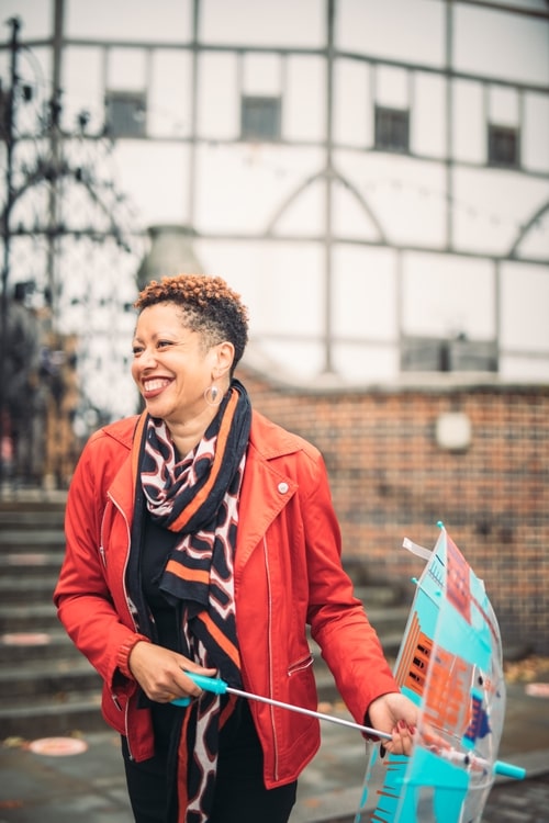 Annette in a bright red jacket laughing and holding an umbrella with the Shakespeare's Globe theatre in the background