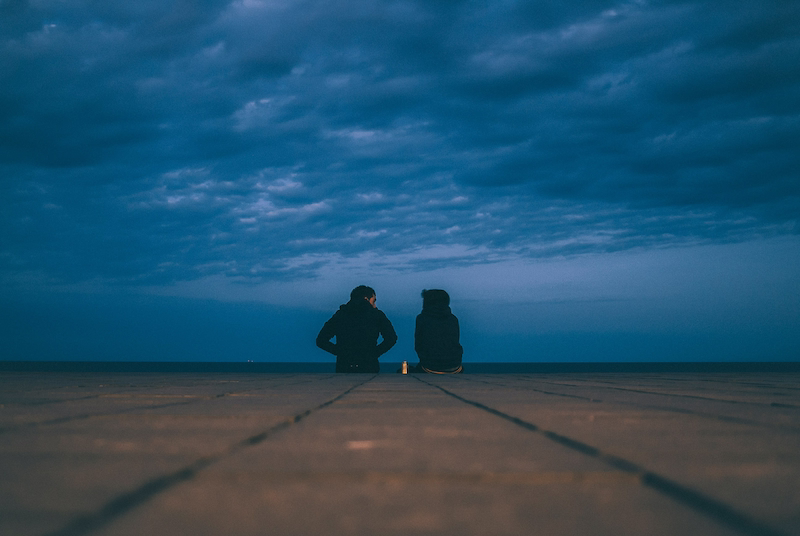 wo figures sitting together looking over the ocean under a cloudy twilight sky, symbolizing teamwork, client trust, and shared vision.