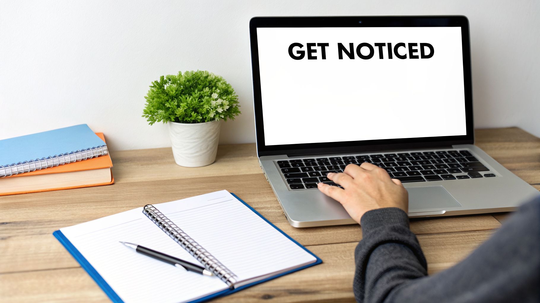 Person typing 'GET NOTICED' on a laptop with books, a notebook, and a plant on a wooden desk.