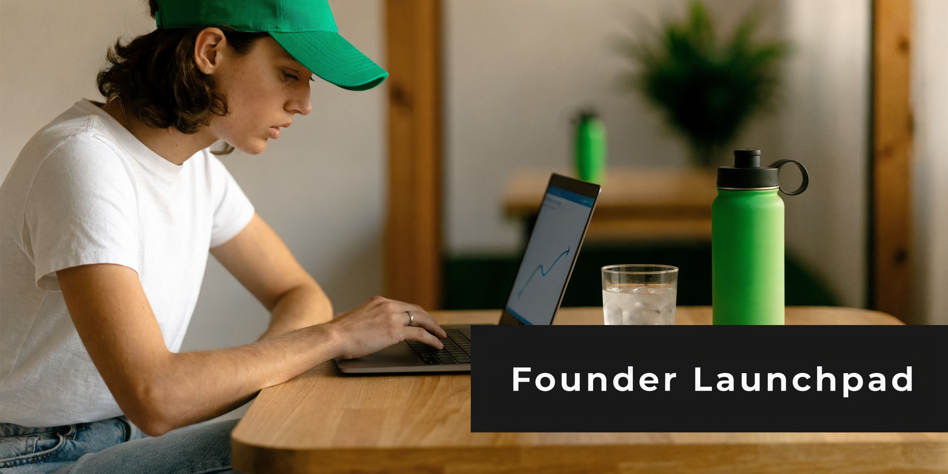 A young person wearing a green cap works on a laptop showing a graph at a wooden table.