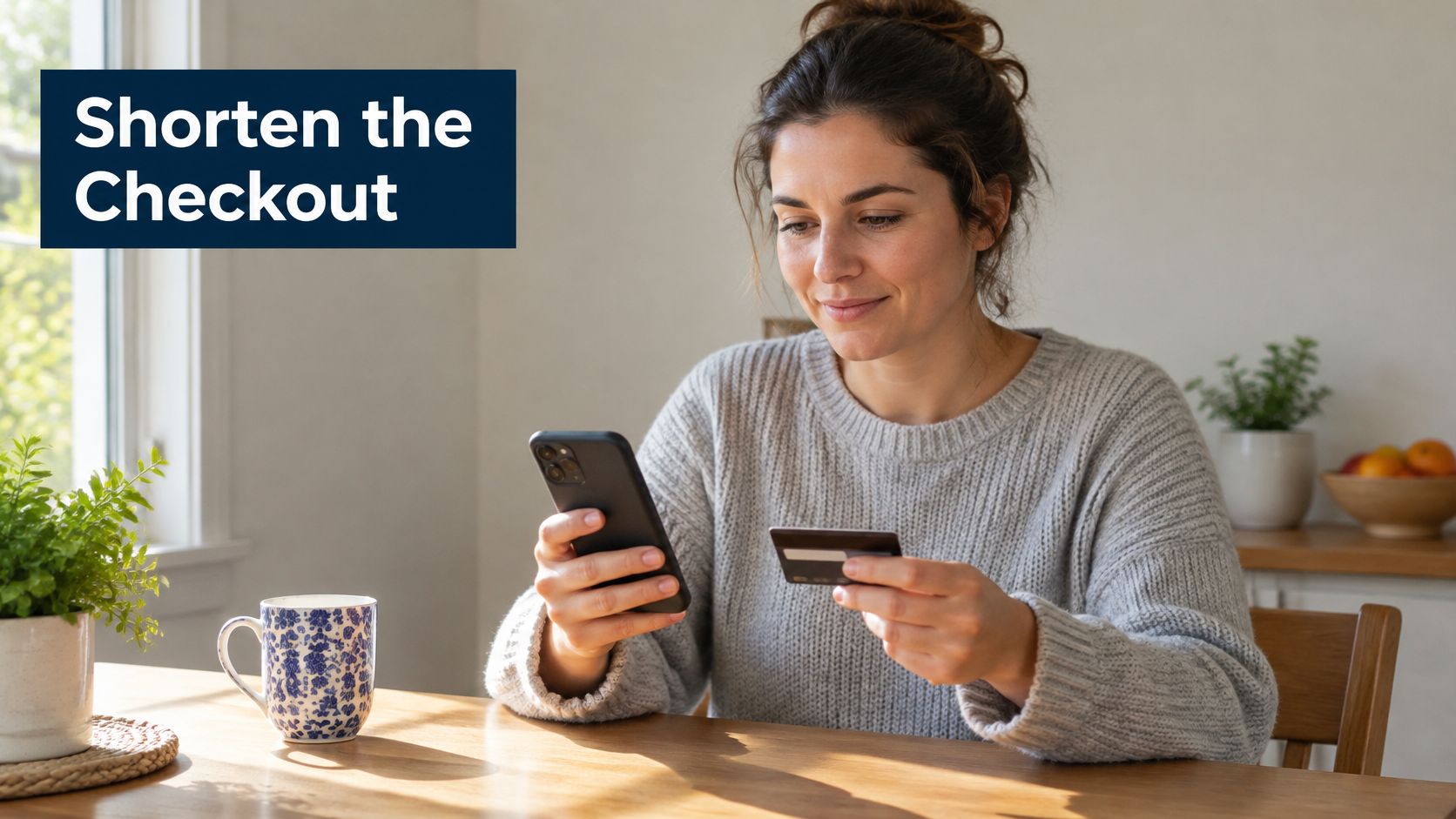 A woman sits at a wooden table using her smartphone and credit card for online shopping.