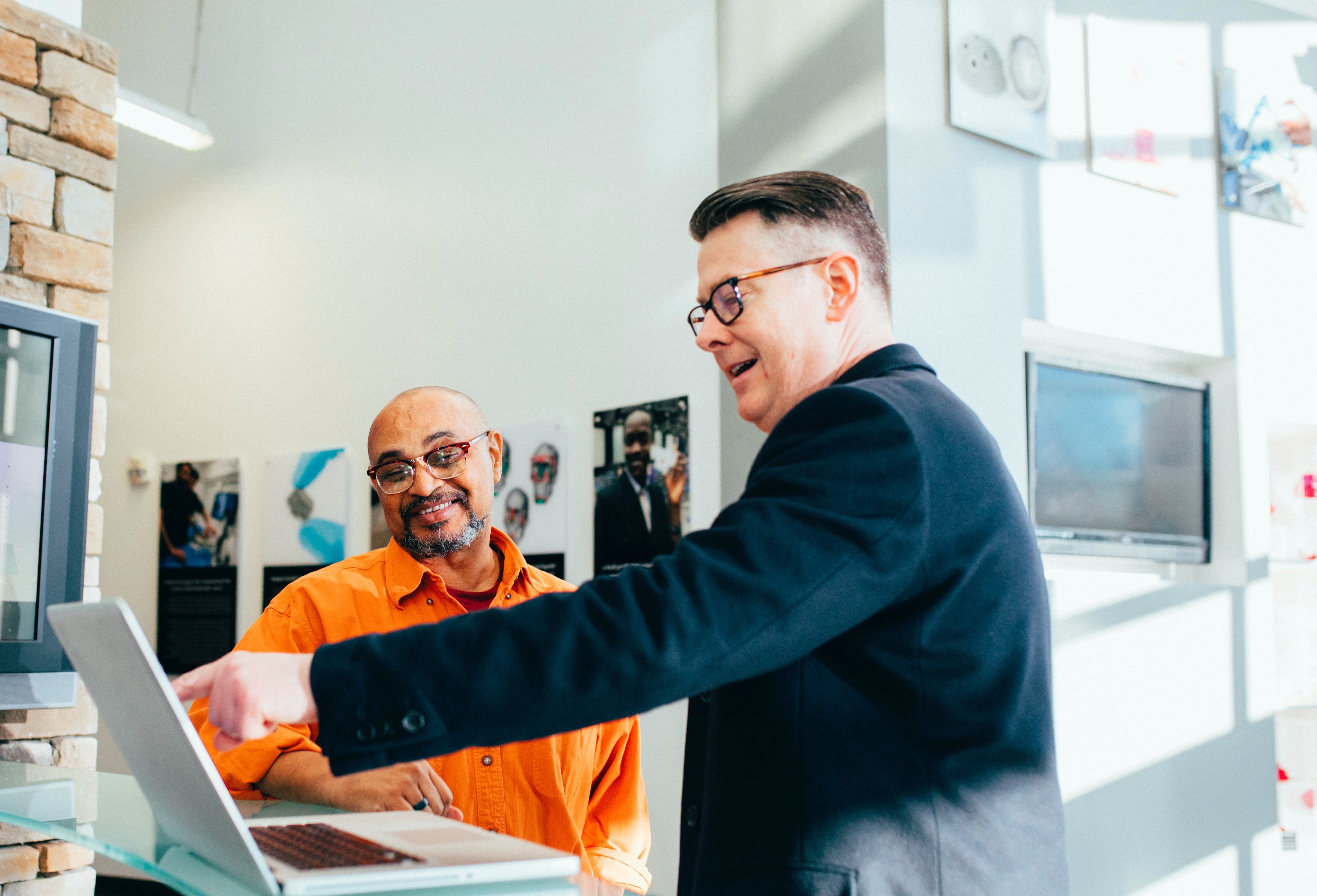 Zwei Personen stehen  für einem Bildschrim. Die Person im Vordergrund zeigt auf diesen. DIe andere Person im Hintergrund folgt mit ihrem Blick der Hand der ersten Person. Beide haben einen Positiven Gesichtsausdruck.
