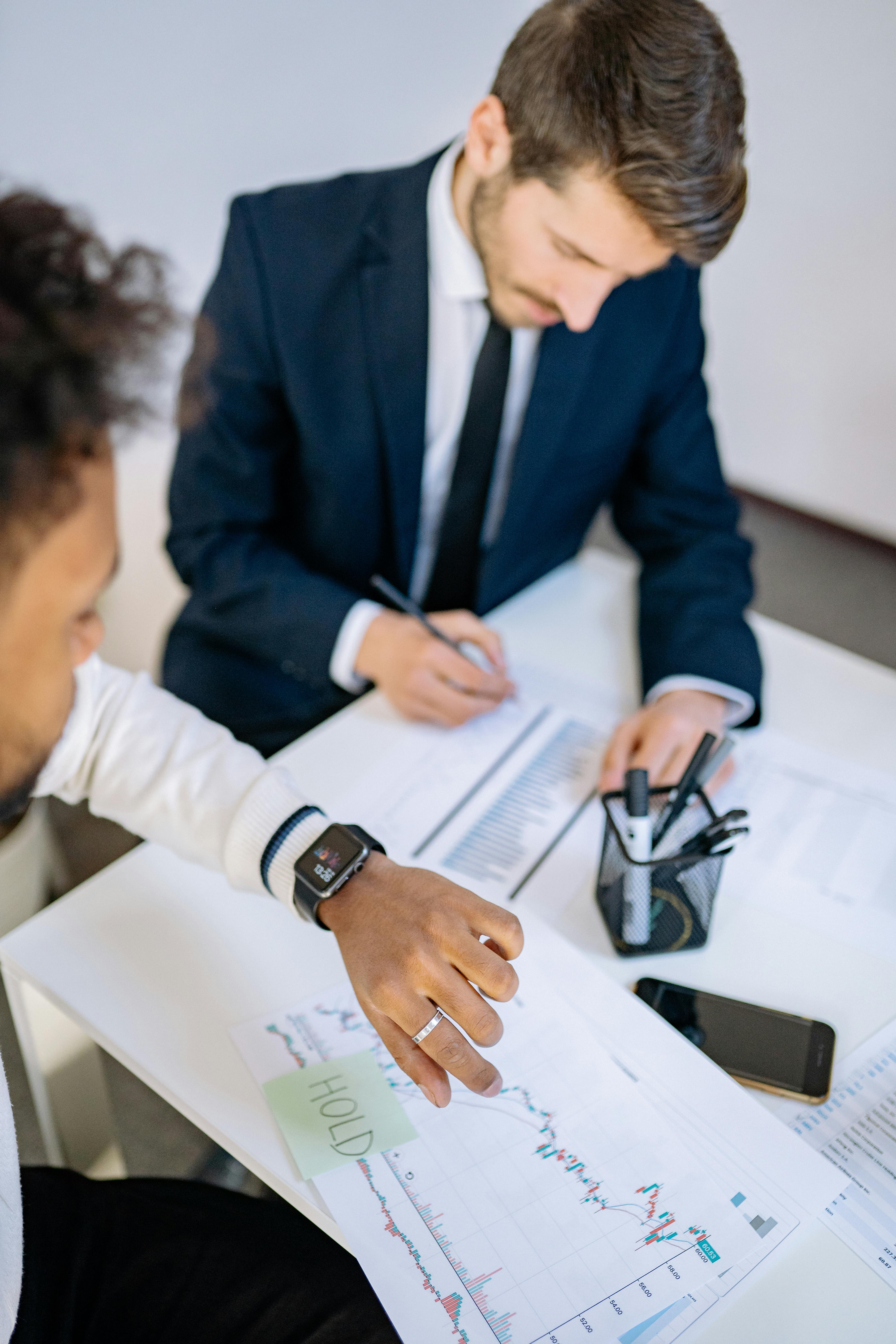 Zwei Personen in Business-Kleidung sitzen gemeinsam am Tisch. Die vordere Person wirft einen Blick auf die Uhr, die hintere Person schreibt mit einem Stift in eine Tabelle auf einem Bogen Papier.