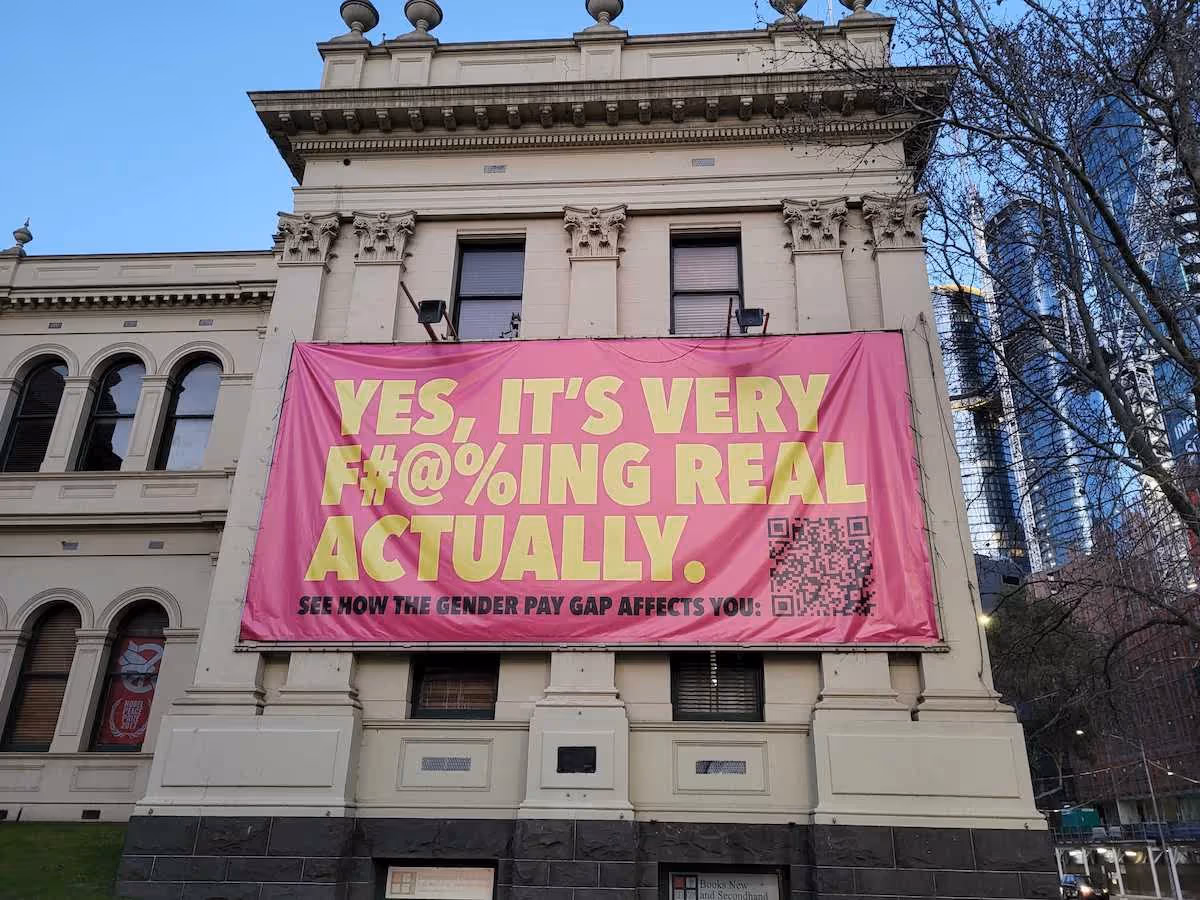 A banner with a QR code is seeing hanging from a building. The banner states "Yes, it's very F-ing real actually. See how the gender pay gap affects you."