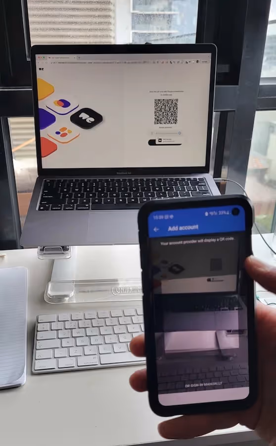 The image shows someone in front of a laptop with a cell phone in their hand. The phone has the Microsoft Authenticator App open and is scanning a QR code on the screen.