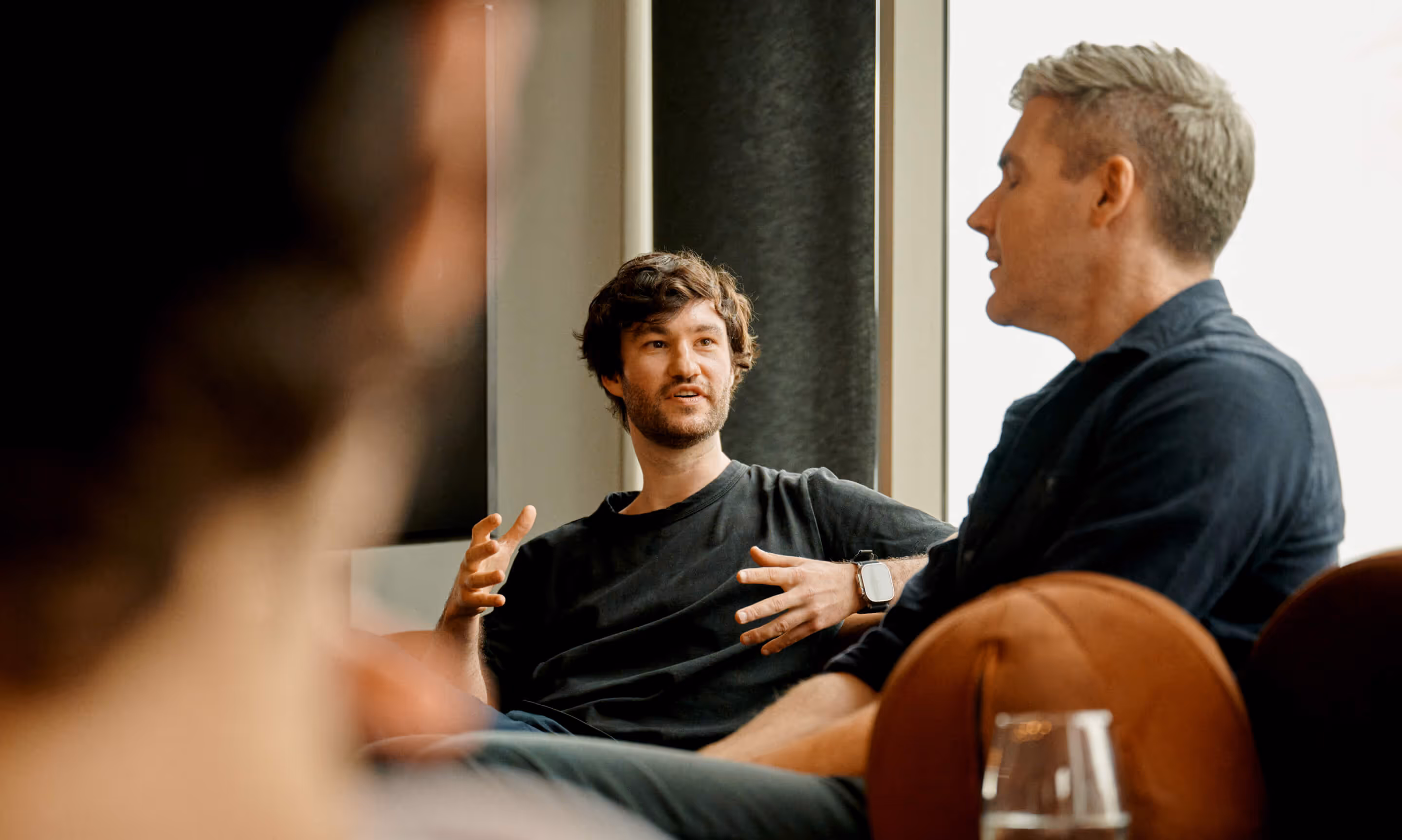Two men engaged in a conversation while sitting indoors, one gesturing with his hands.