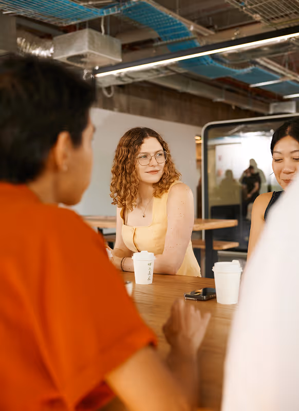 Three people sitting around a wooden table in a modern office space, with coffee cups and a smartphone on the table.