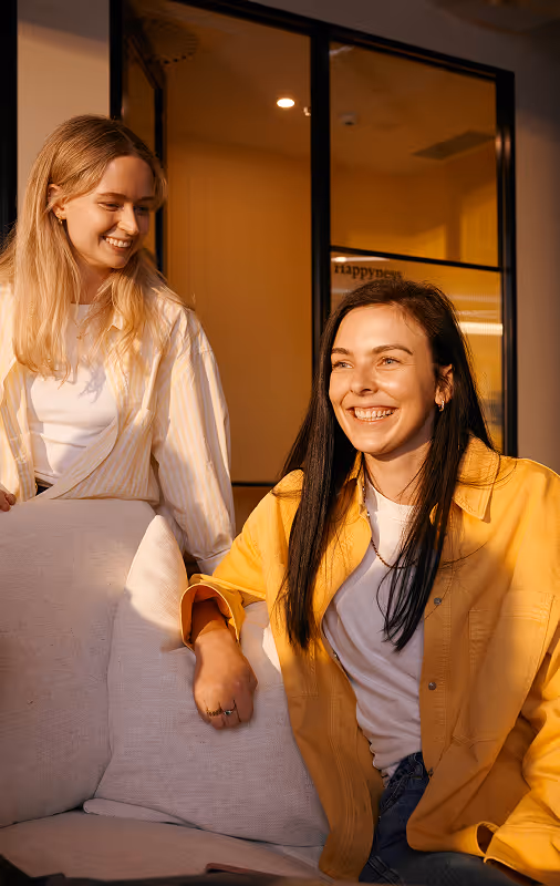 Two women smiling indoors, one in a yellow jacket seated on a beige couch and the other standing beside her in a light striped shirt.