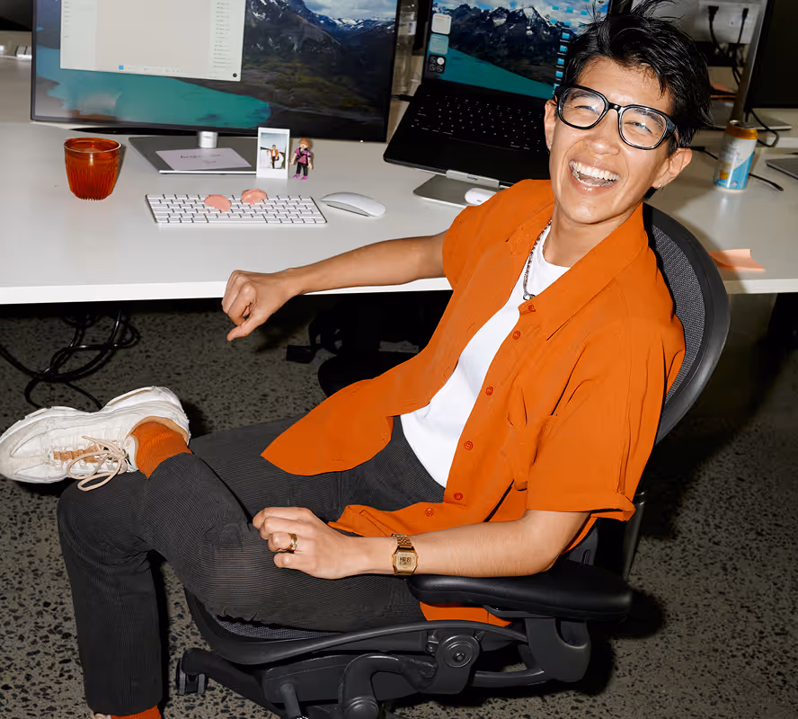 Smiling person wearing glasses and an orange shirt sitting casually in an office chair with crossed legs in front of a desk with computer monitors.