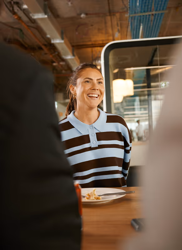 Smiling woman wearing a blue and brown striped shirt sitting at a table in a modern office setting.