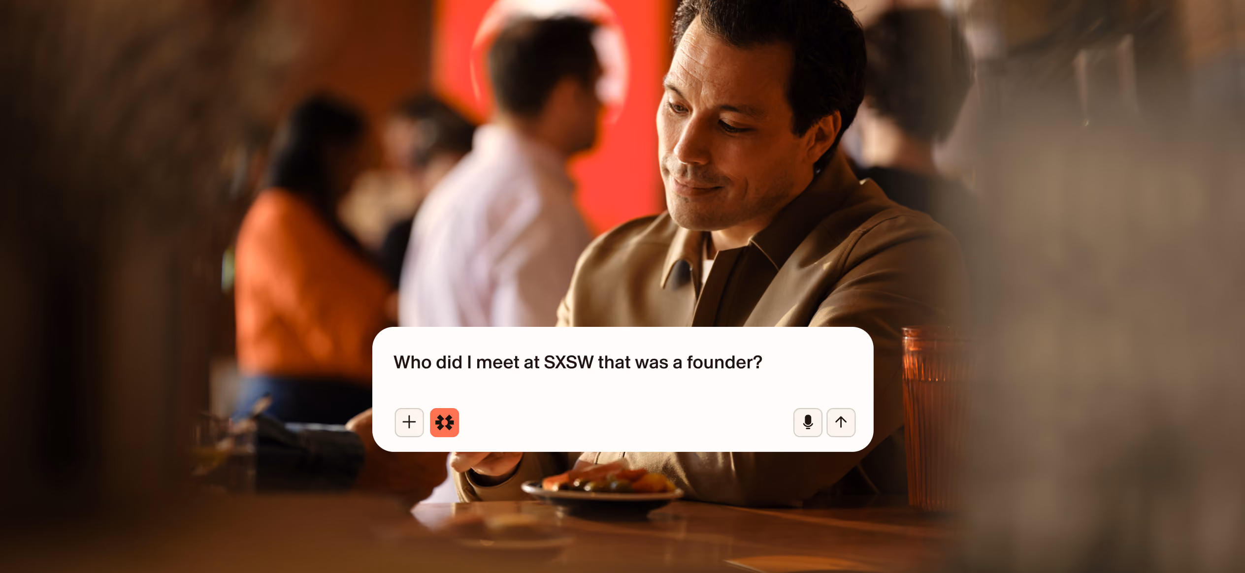 Man sitting at a restaurant table looking at his phone with a displayed text message that reads, 'Who did I meet at SXSW that was a founder?' Other people are blurred in the background.