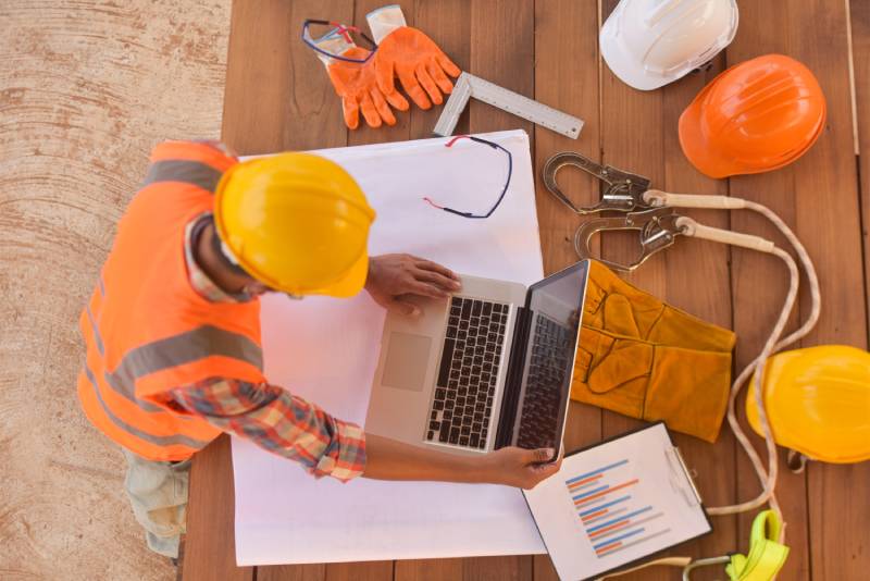 Construction worker with laptop, blueprints, and safety gear.