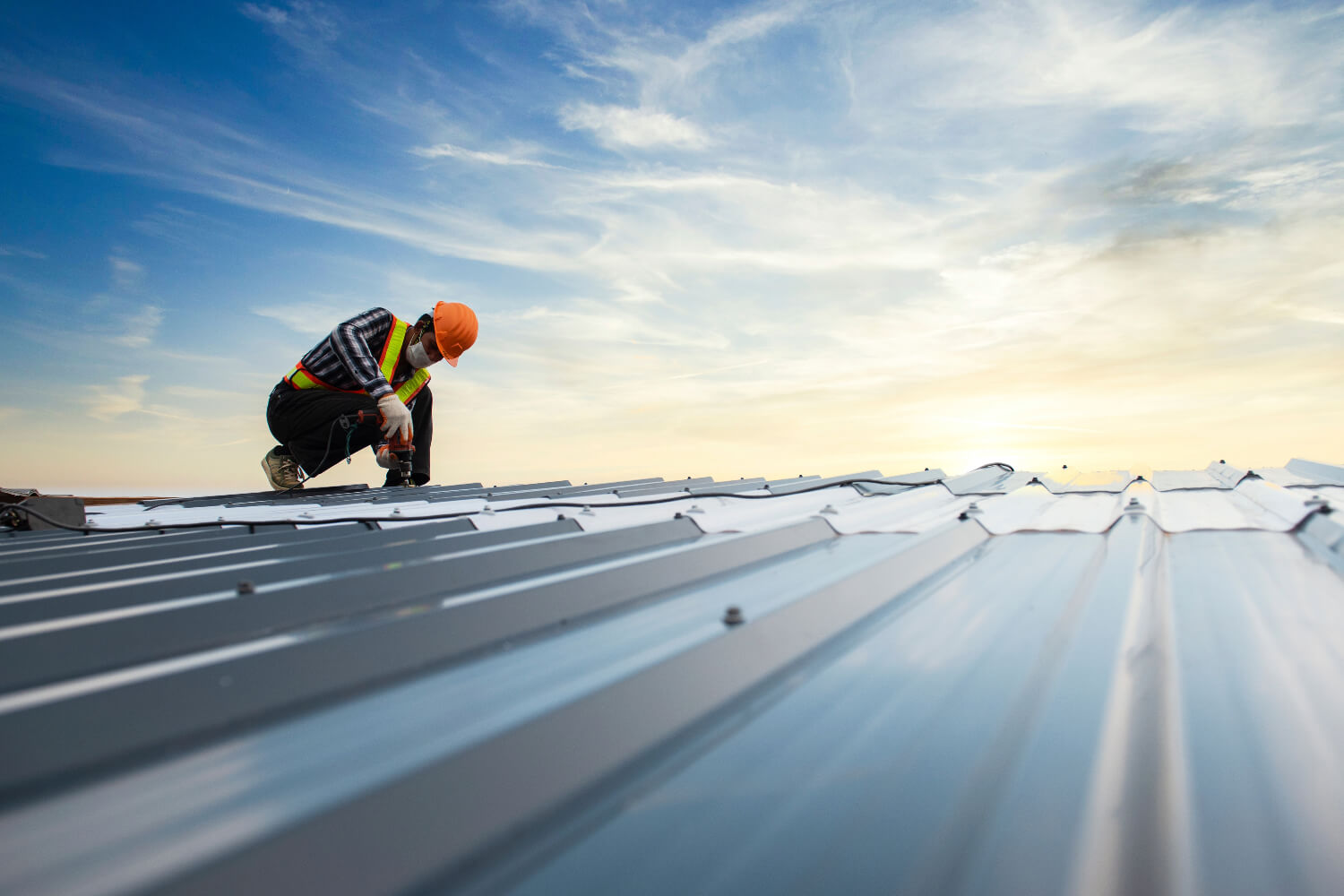 Roofer in safety gear working on metal roof at sunset.