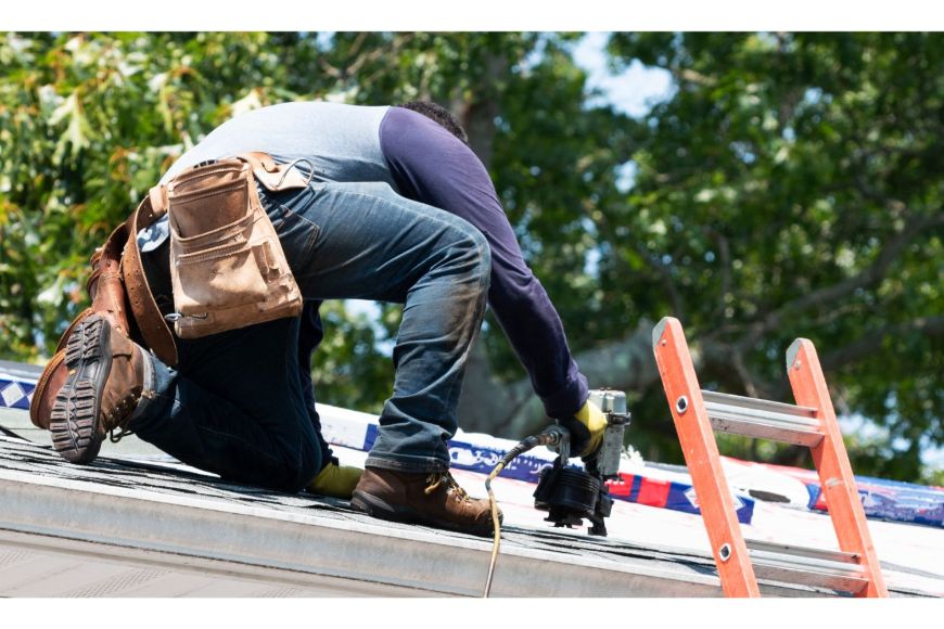 Roofer with toolbelt using nail gun on gray shingles near red ladder.