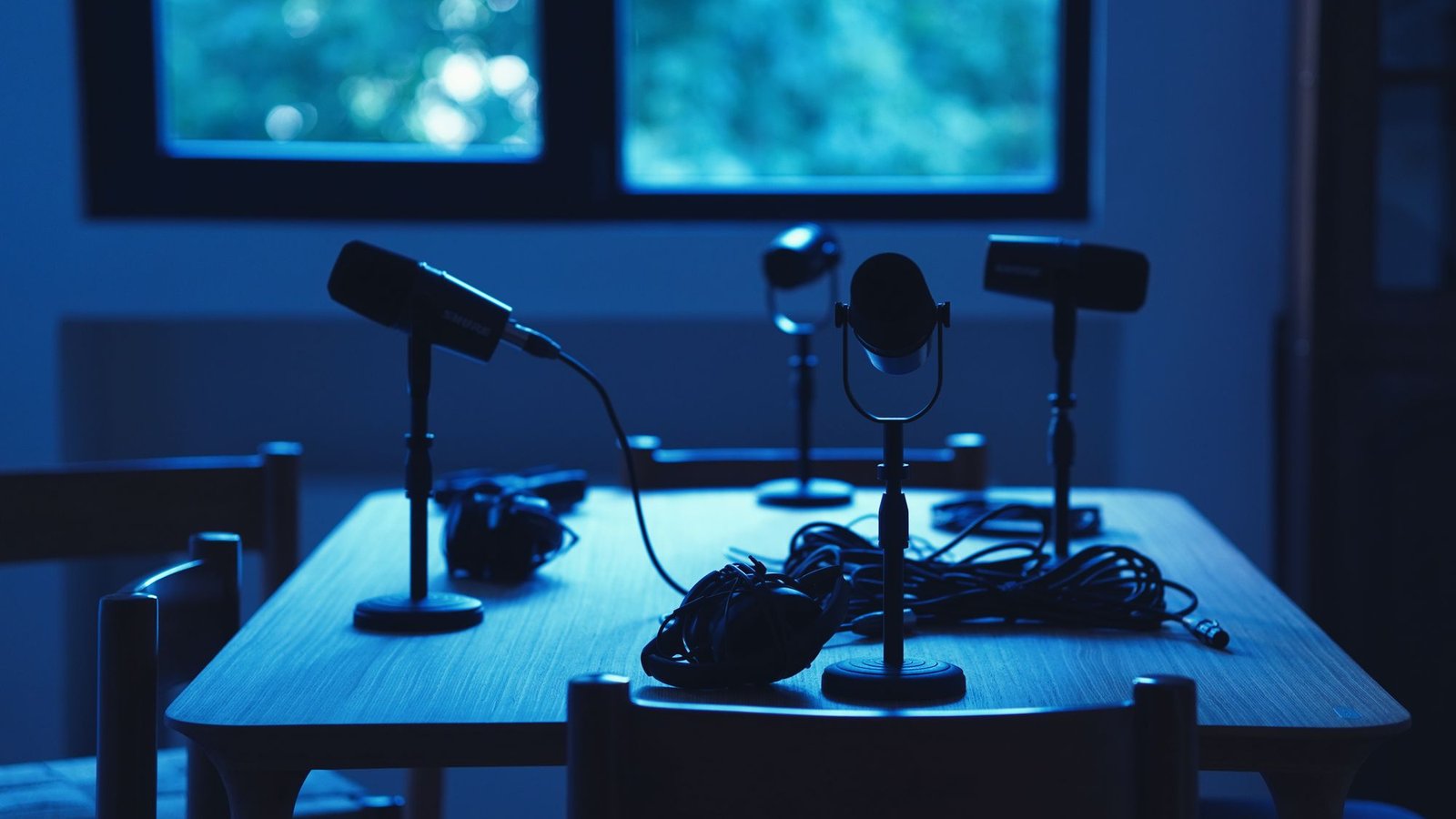 Podcast microphones on desk stands around a wooden table in a dimly lit, blue-toned recording room