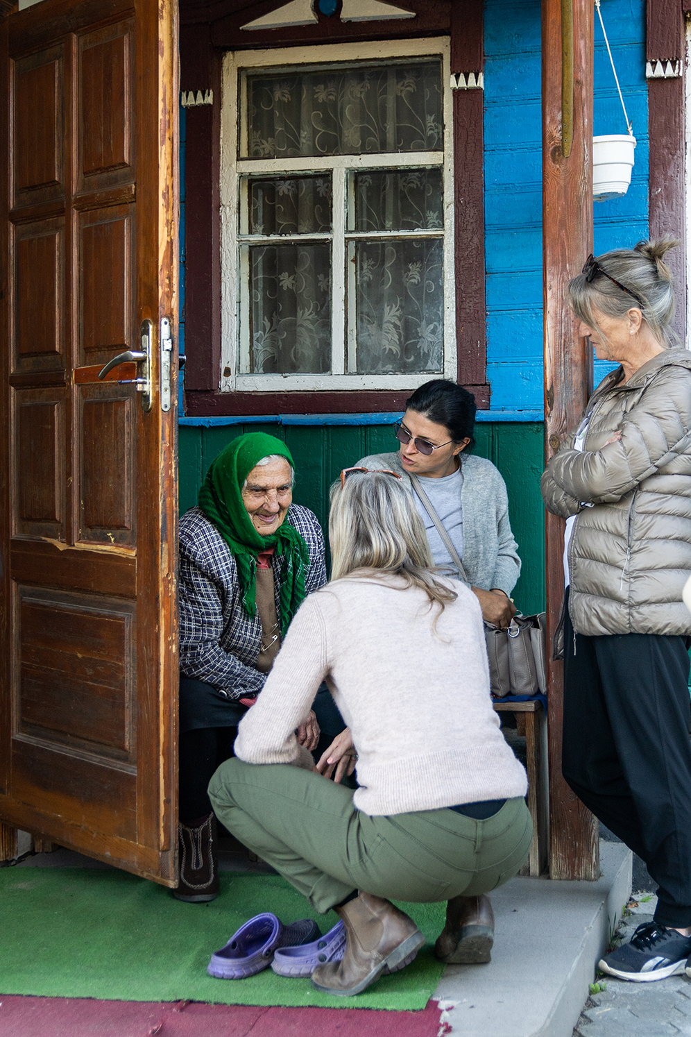 Three women visiting an elderly woman seated at the entrance of a blue wooden house with a green door frame.
