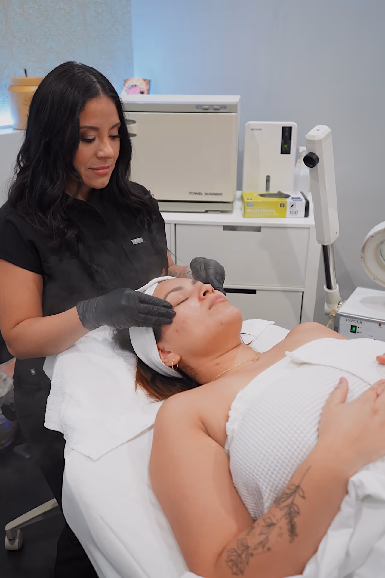 A professional esthetician at Refine Skin Kare performing a facial treatment on a client. The esthetician, wearing a black uniform and gloves, gently presses on the client's face, who is lying on a treatment bed wrapped in a white towel. The room features modern equipment, including a vaporizer and a towel warmer, with a clean and serene ambiance. The client has a floral tattoo on their arm, adding a personal touch to the scene.